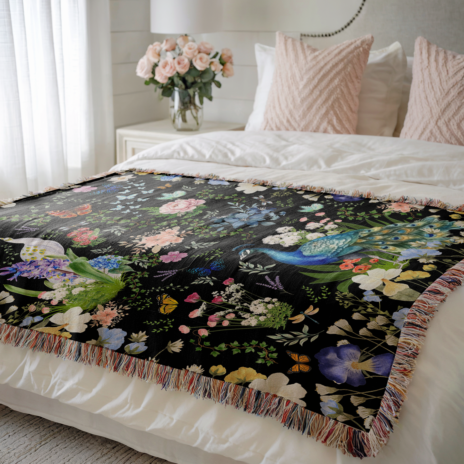 Black Peacock Floral Woven Blanket with Fringes on a bed with decorative pillows and flowers in the background.