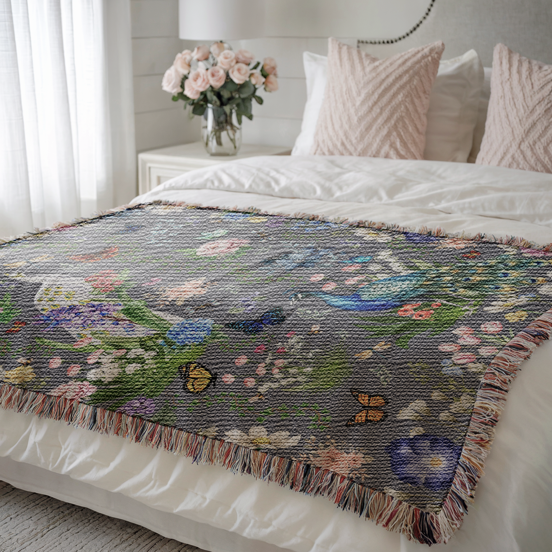 Dark Gray Peacock Floral Woven Blanket with Fringes on a bed with pink pillows and flowers in the background.