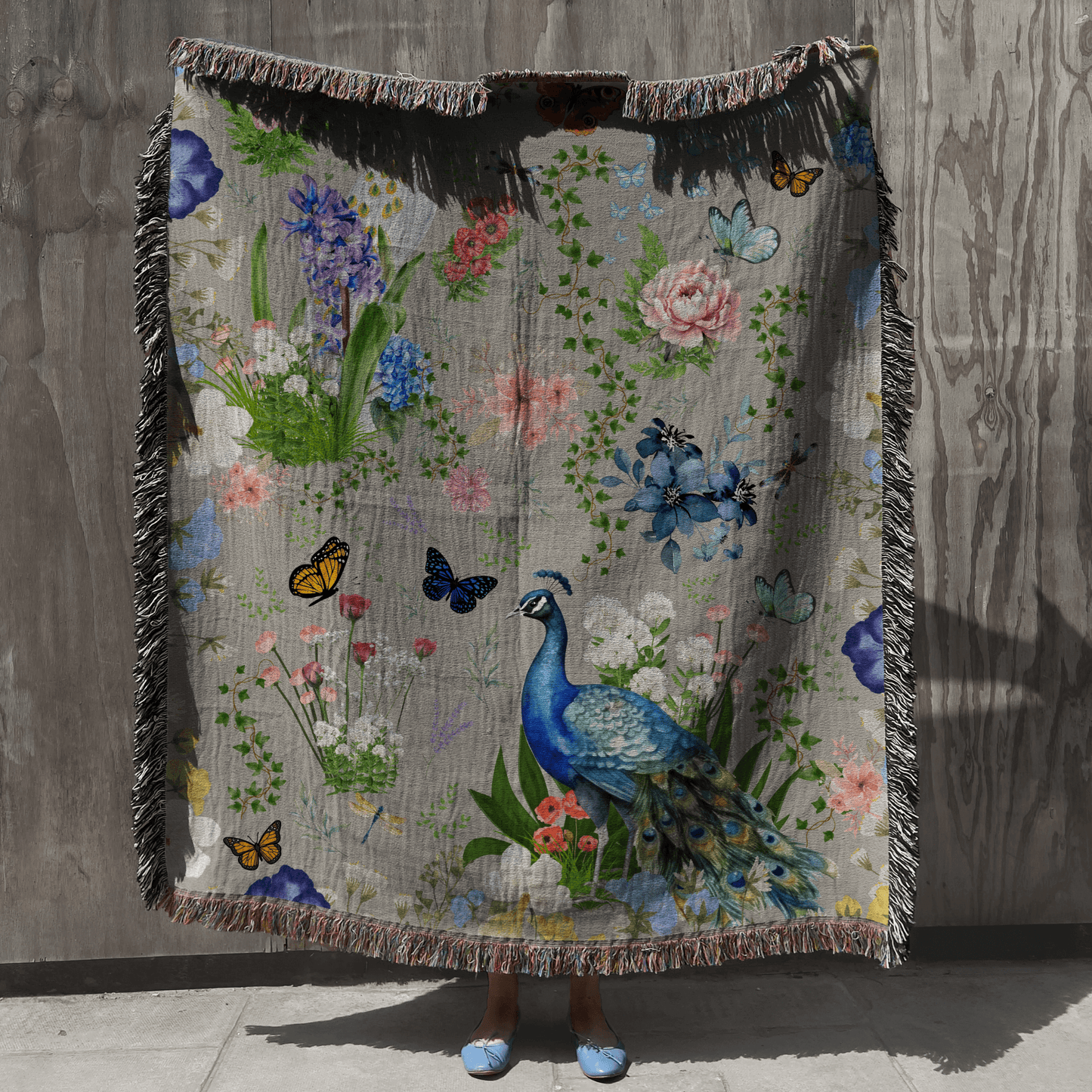 A person holding up the Gray Peacock Floral Woven Blanket with Fringes against a gray wood wall.