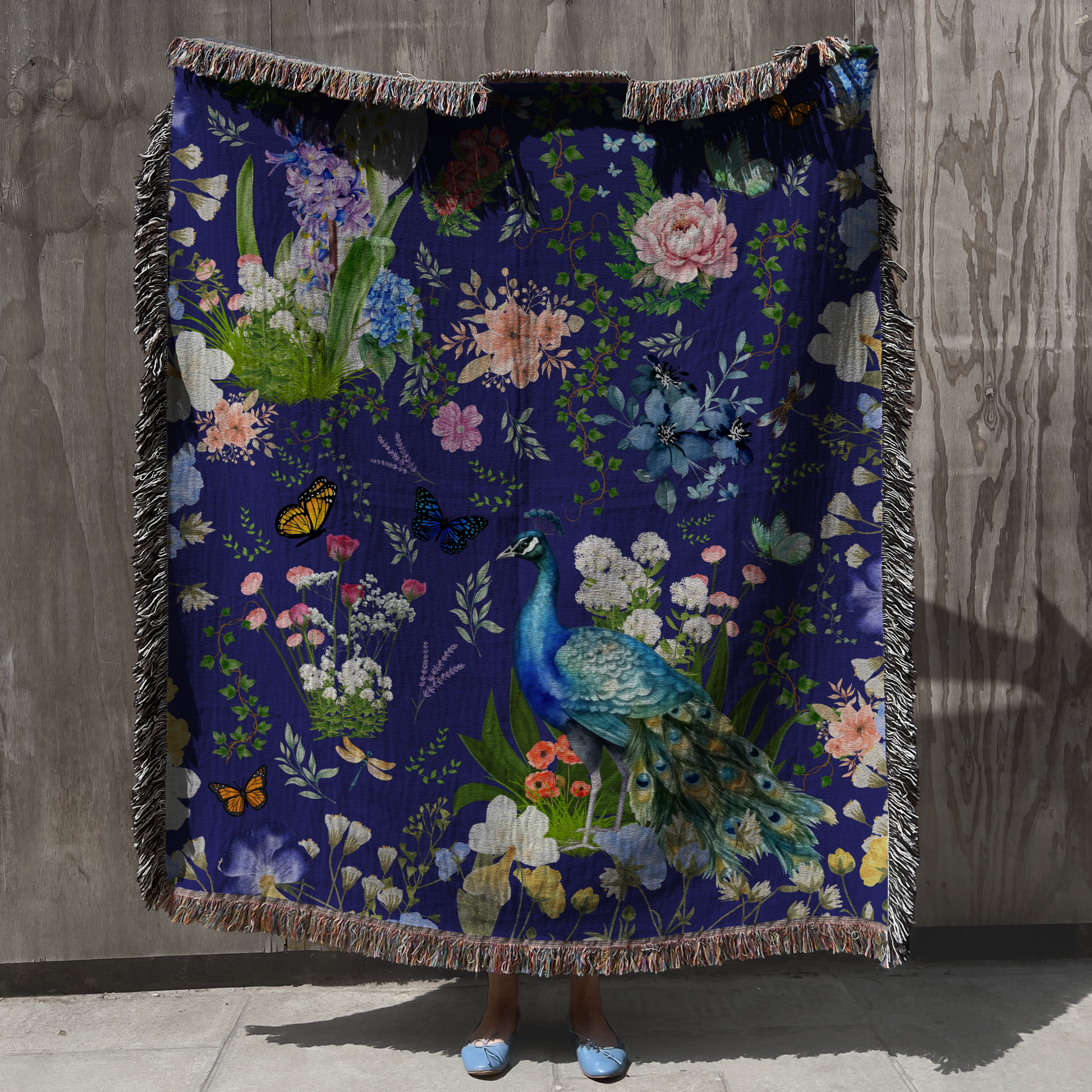 Person holding up the Blue Peacock Floral Woven Blanket with Fringes against a gray wall 