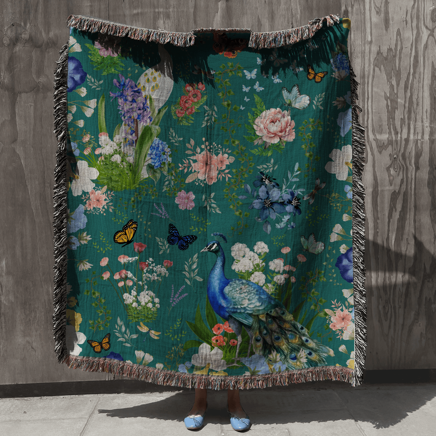 Teal Peacock Floral Woven Blanket with Fringes held up by a person against a wooden wall.