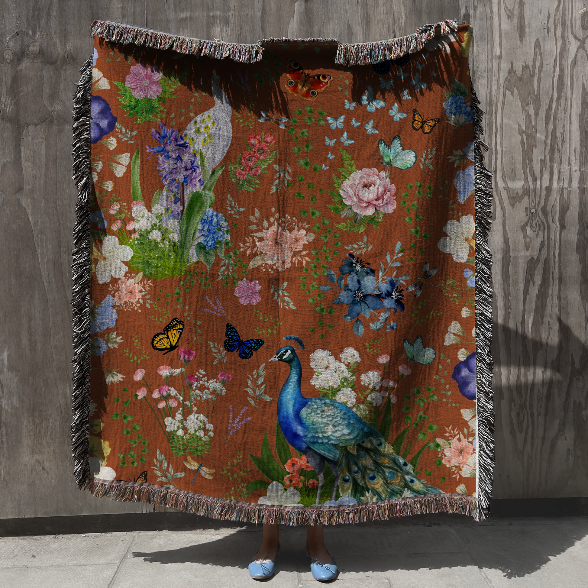 A person holding up the Cinnamon Brown Peacock Floral Woven Blanket against a gray wooden panel background